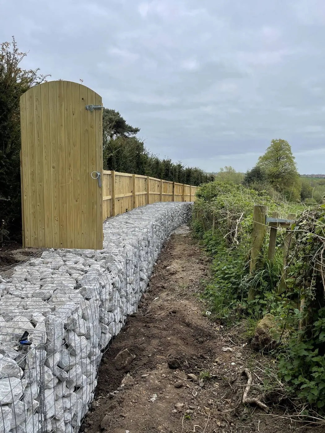 A long gabion wall of white stone running beside a new timber gate and fence along a hedge line