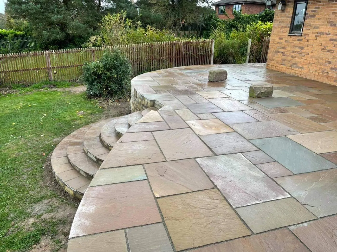A curved sandstone terrace with wrap-around steps down to a lawn, beside a chestnut paling fence