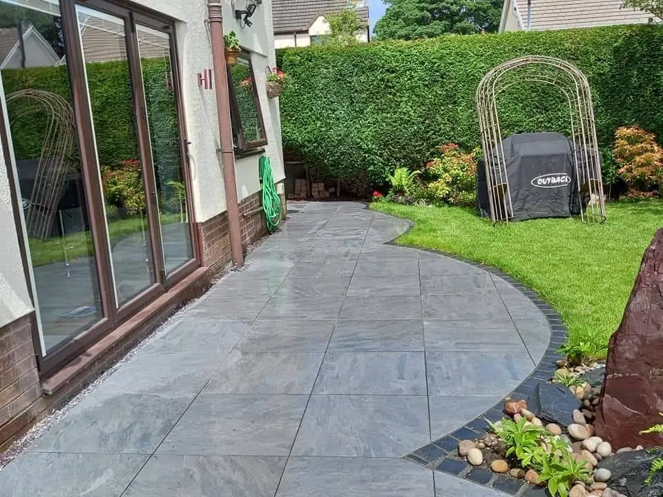 A porcelain path running along the side of a house to a curved patio edge, with a rose arch beyond