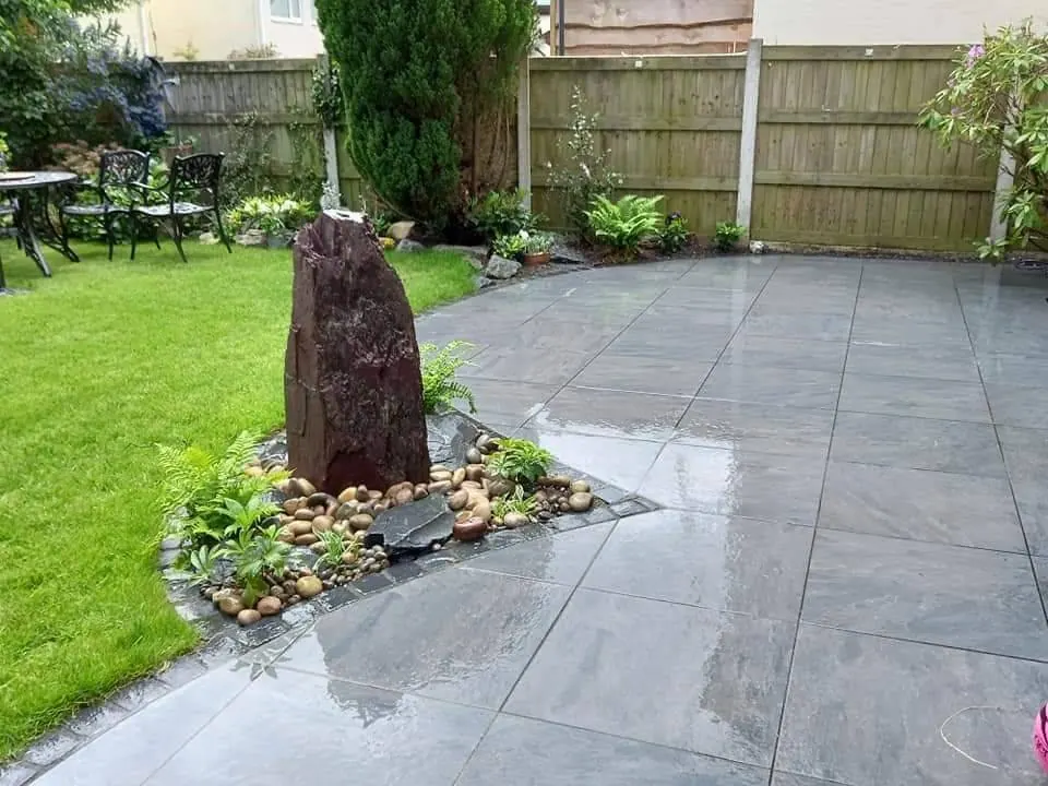 A grey porcelain patio wrapping around a standing stone set in pebbles and ferns, beside a lawn