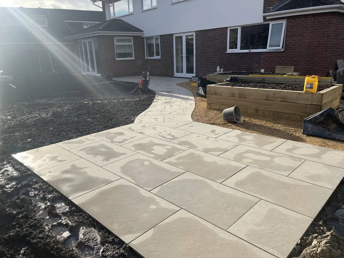 A new pale stone terrace curving away from the house, with timber sleeper raised beds being built alongside