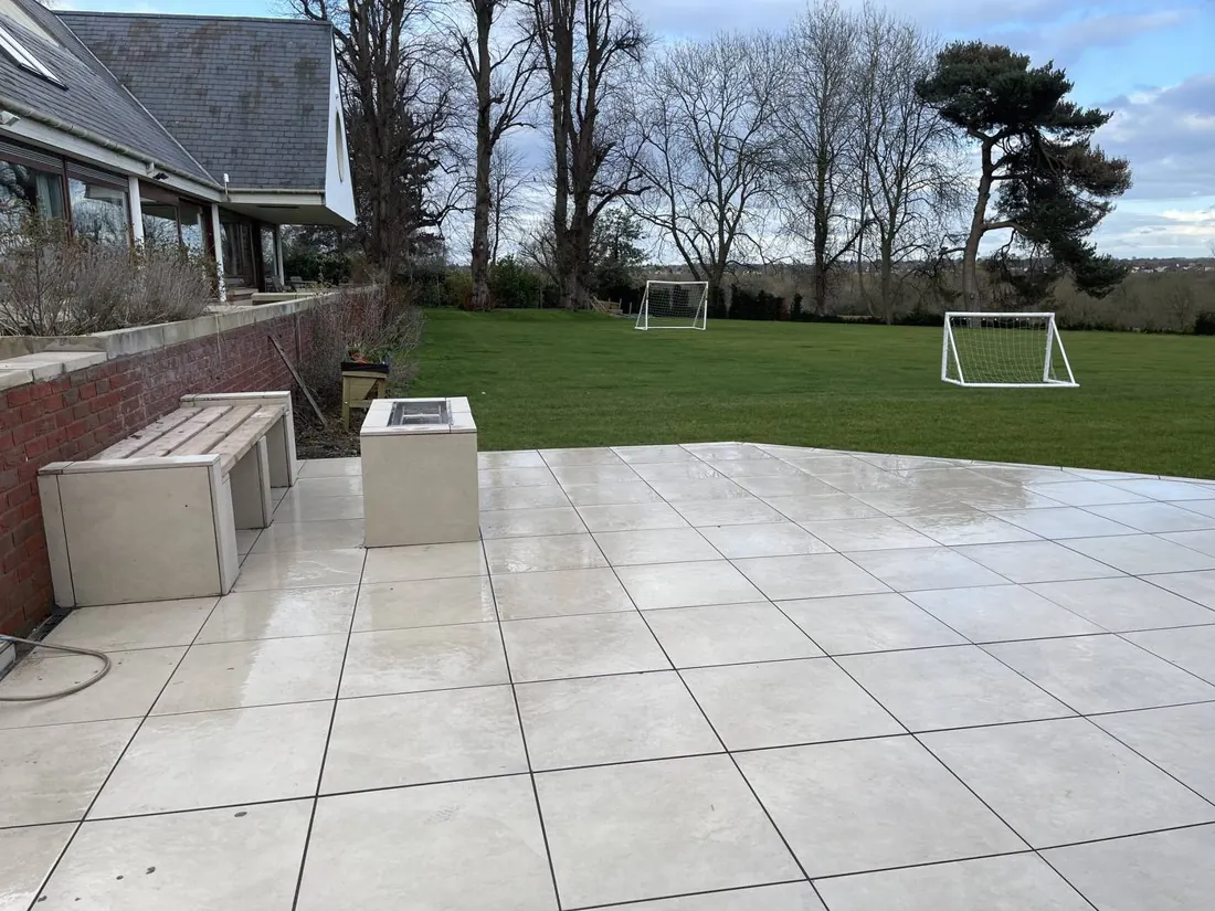 A large porcelain terrace meeting an open lawn, with a bench and fire table against a brick wall